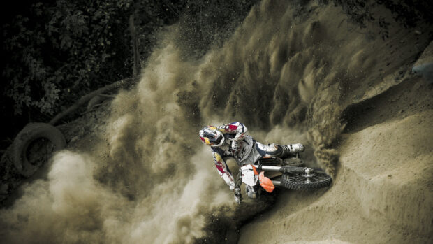 A rider on a KTM dirt bike kicks up dust while making a sharp turn on a dirt track