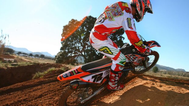 A rider in red and white gear riding a KTM dirt bike on a dirt track under a clear blue sky