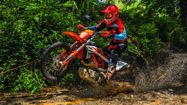 A rider in red gear expertly rides a KTM dirt bike through a muddy forest trail