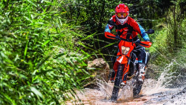 A rider on a KTM dirt bike splashes through a muddy trail surrounded by green foliage