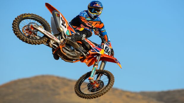 A rider performing a jump on an orange KTM dirt bike under a clear blue sky