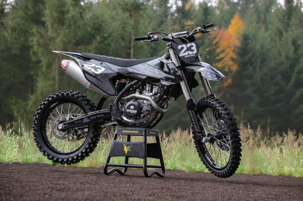 A black and gray KTM dirt bike stands on a stand in a forest setting