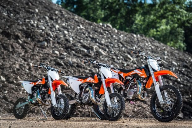 Three orange and white KTM dirt bikes are lined up on a dirt track with a rocky hill background
