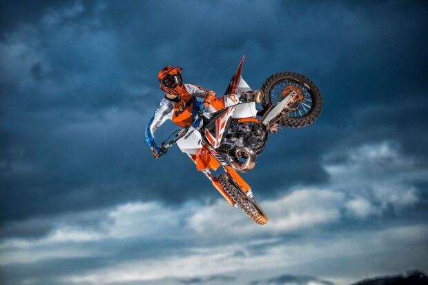 A rider in orange and white gear performs a jump on a KTM dirt bike against a dramatic cloudy sky