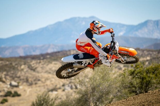 A rider on a KTM dirt bike jumps high over a dirt trail with mountains in the background