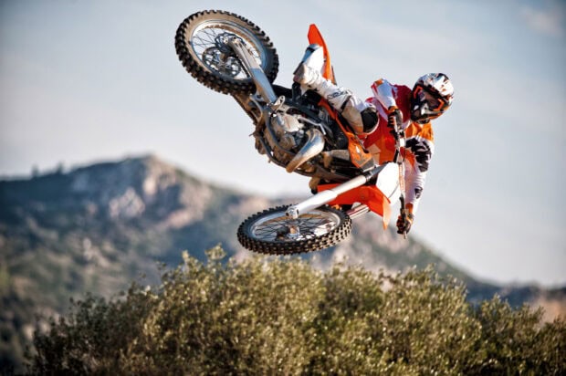 A rider performs an impressive jump on a KTM dirt bike over natural terrain with mountains in the background