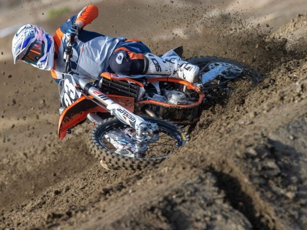 A rider on a KTM dirt bike is skillfully cornering on a muddy dirt track kicking up dust