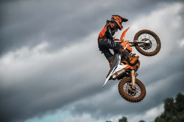 A rider performs a high jump on an orange KTM dirt bike under a cloudy sky