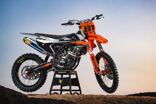 A KTM dirt bike with orange and black design stands on a dirt surface against a clear sky background