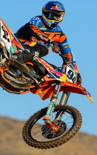 A rider on an orange KTM dirt bike performs a jump against a clear blue sky background