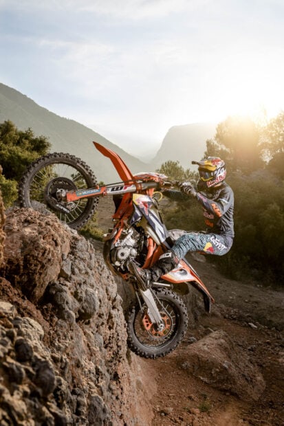 A rider performs a wheelie on an orange KTM dirt bike climbing rocky terrain at sunset