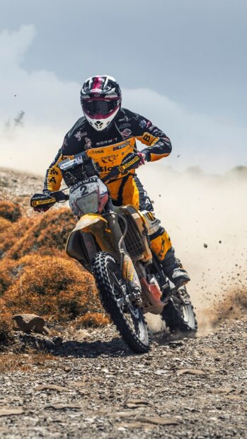 A rider on a KTM dirt bike races through a rocky off road trail kicking up dust
