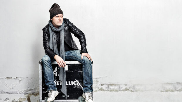 Lars Ulrich is sitting casually on a Metallica equipment case against a white wall, HD Desktop Wallpaper
