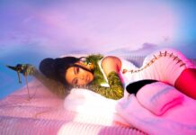 Luxury fashion woman with bold makeup and glamorous outfit lying on pink bedding under colorful sky.