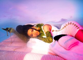 Luxury fashion woman with bold makeup and glamorous outfit lying on pink bedding under colorful sky.
