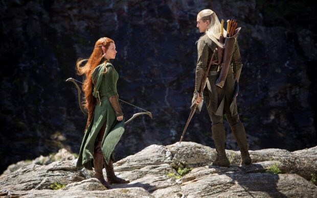 Legolas standing on rocky terrain with an elf companion in green attire and bows in hand