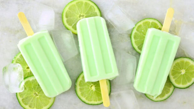 Three lime flavored popsicles on ice and lime slices arranged on a surface