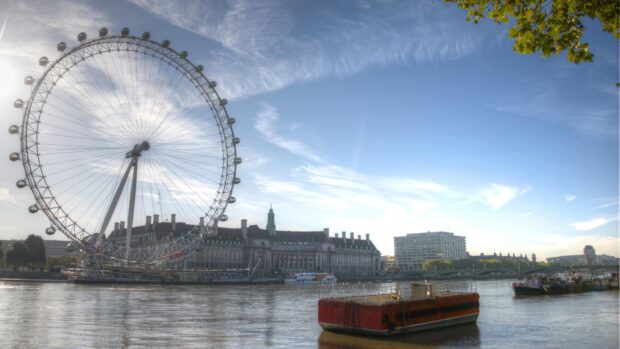 The London Eye stands tall above the River Thames with buildings and boats alongside under a blue sky, 4K Desktop Wallpaper
