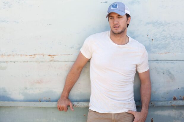 Luke Bryan is leaning against a rustic wall wearing a white t shirt and cap, 2K Desktop Wallpaper