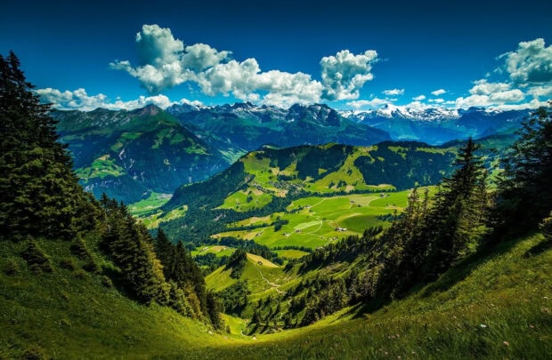 A panoramic view of green hills with scattered houses and towering mountains, HD Desktop Wallpaper