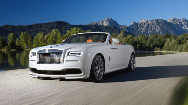 A luxury Rolls Royce Dawn drives along a scenic mountain road under a clear blue sky, 2K Desktop Wallpaper