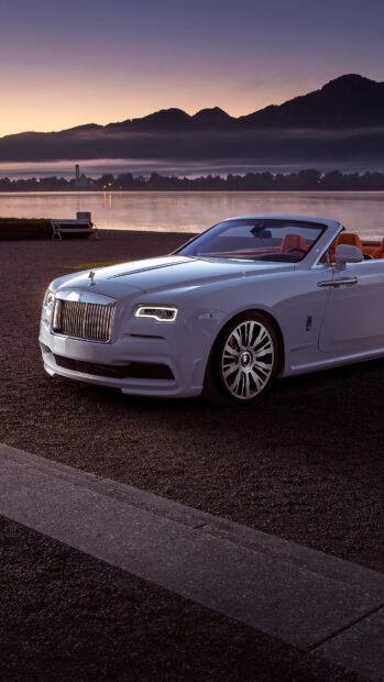 A white Rolls Royce Dawn is parked near a calm lake with mountain silhouettes in the background at dusk, 4K Mobile Wallpaper