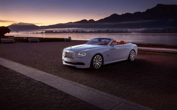 Luxury Rolls Royce Dawn is shown parked by the water with mountains and sunset in the background, 2K Desktop Wallpaper