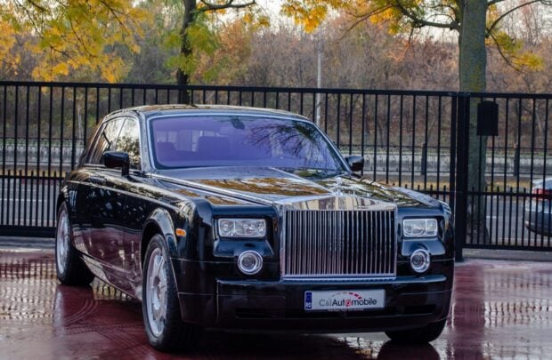 A black Rolls Royce Phantom is parked near a metal fence with autumn trees in the background, 2K Desktop Wallpaper