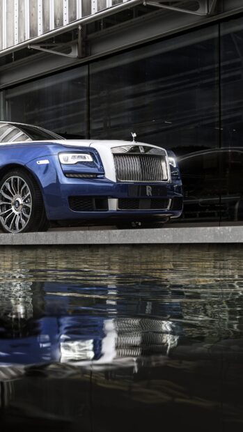 A blue and white Rolls Royce Ghost parked near reflective water under modern architecture, 4K Phone Wallpaper