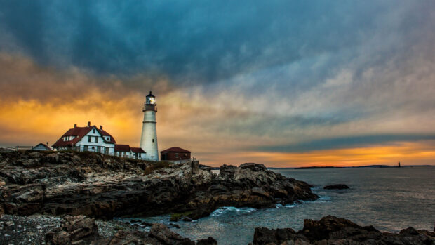 A Maine lighthouse stands on rocky cliffs by the ocean during a colorful sunset with vibrant sky, 4K Desktop Wallpaper