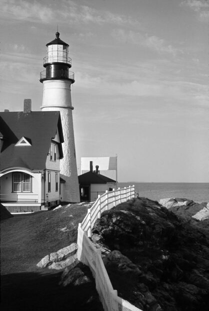 A lighthouse stands near a rocky coast in Maine with surrounding buildings and a fence, HD Mobile Wallpaper