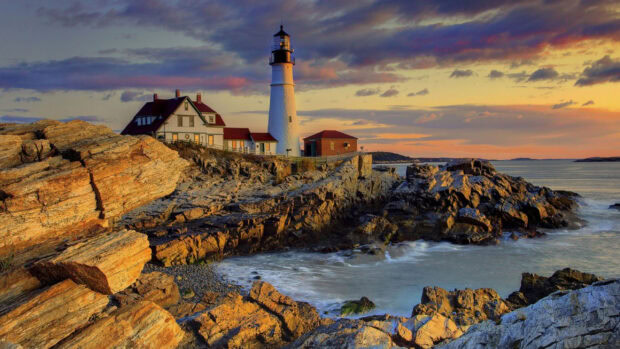 The image shows Maine with a lighthouse on the rocky coastline during sunset, 2K Desktop Wallpaper