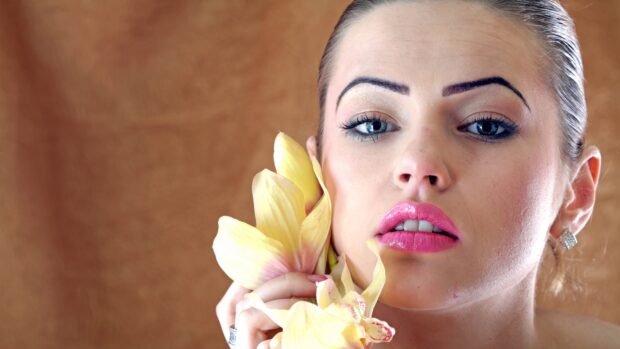 A woman with vibrant makeup holding yellow orchid flowers near her face with elegant eye makeup