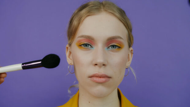 Woman with colorful makeup applying cosmetics with brush on purple background