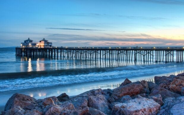 The Malibu pier extends over calm ocean waves at sunset with rocky shore in the foreground
