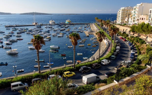 A scenic view of Malta harbor with numerous boats and palm trees along the coastal road