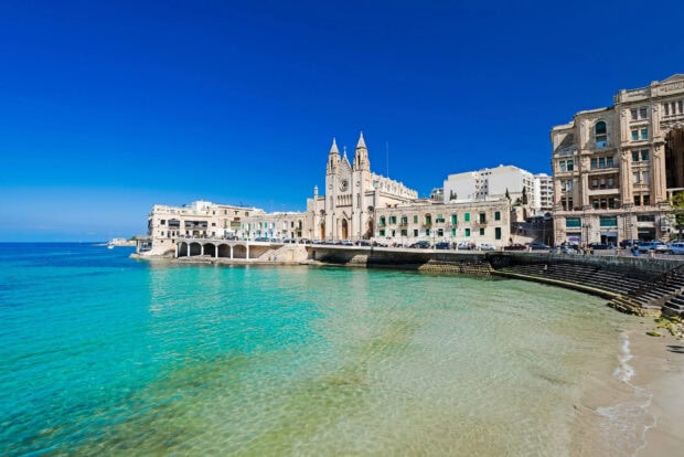 Clear turquoise waters next to historic Malta buildings under a bright blue sky