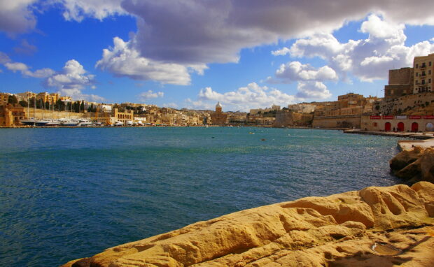 View of Malta coastline with historic buildings under cloudy blue sky and calm sea water