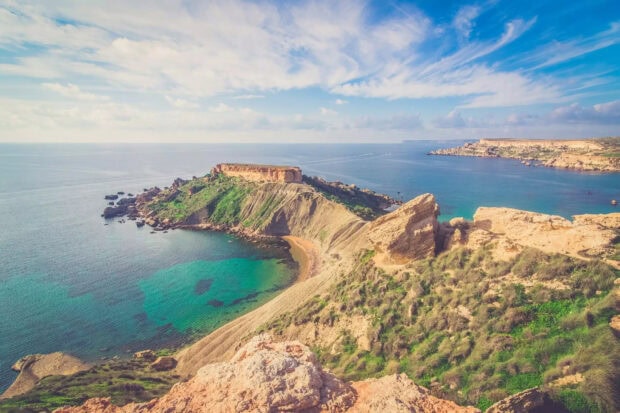A scenic coastal landscape of Malta with rocky cliffs and clear turquoise waters under a partly cloudy sky