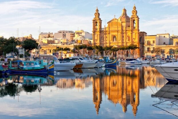 A picturesque view of Malta harbor with boats and historic buildings reflected in calm water during sunset