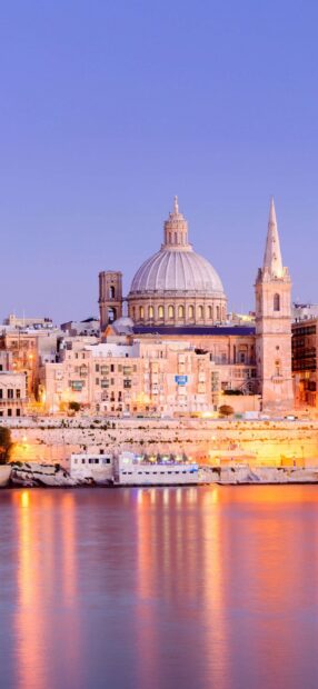 The skyline of Malta features historic architecture with domes and spires reflecting in calm water at sunset