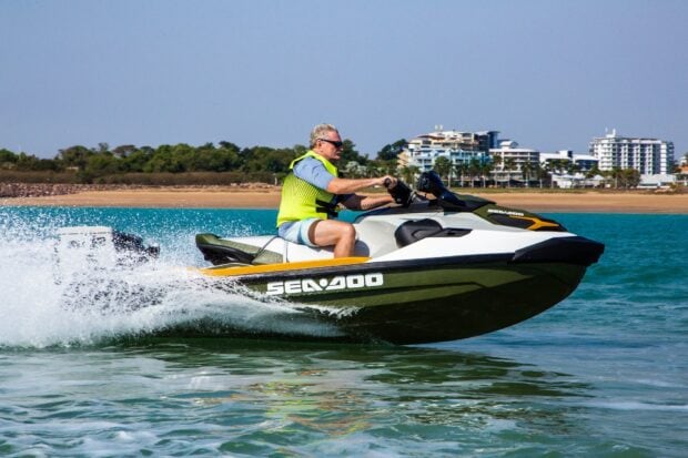 A man wearing a bright vest is riding a Sea Doo jet ski on calm water near the shore, HD Desktop Wallpaper