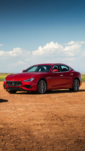 Red Maserati car parked on dry ground under a clear blue sky with clouds