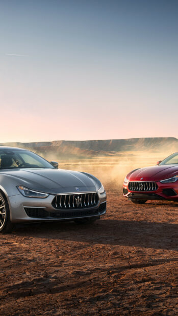 Two Maserati cars parked on dry land with mountain background at sunset