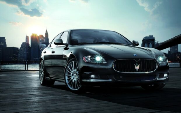 A sleek black Maserati is parked on a wooden pier with city skyline in the background at sunset
