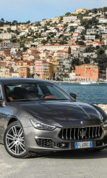 Grey Maserati car parked near the sea with colorful houses on the hillside in the background