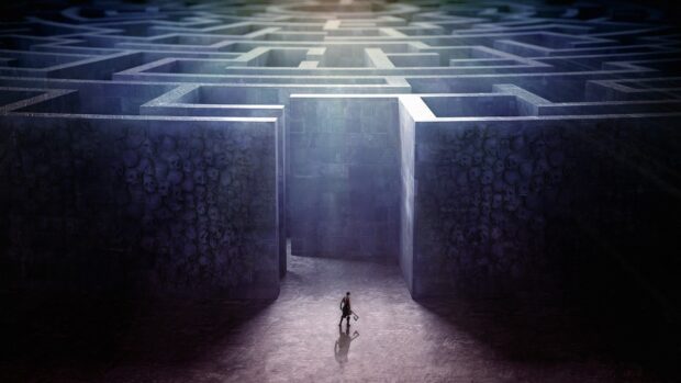 A lone man stands before an enormous maze with towering walls covered in skull carvings