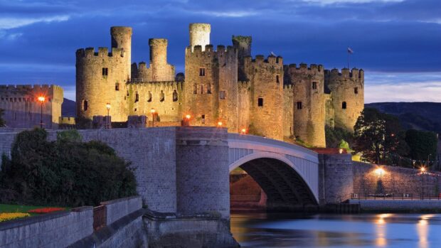 A medieval castle in Wales stands illuminated near the water at dusk under a deep blue sky, HD Desktop Wallpaper