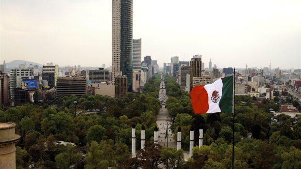 Mexico City Wallpapers show a flag over the city with tall buildings and green trees, 2K Desktop Wallpaper