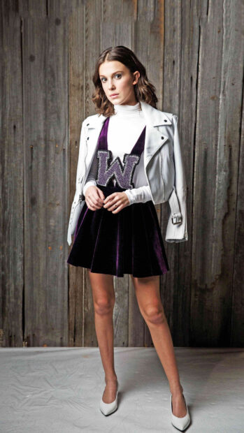 Millie Bobby Brown stands wearing a white jacket and purple skirt in front of a wooden wall, 4K Phone Wallpaper